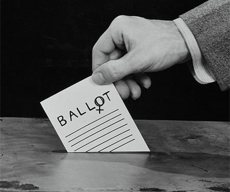 A black-and-white photo of a hand placing a ballot (where the 'o' is the symbol for Venus, which is associated with women) into a ballot box.